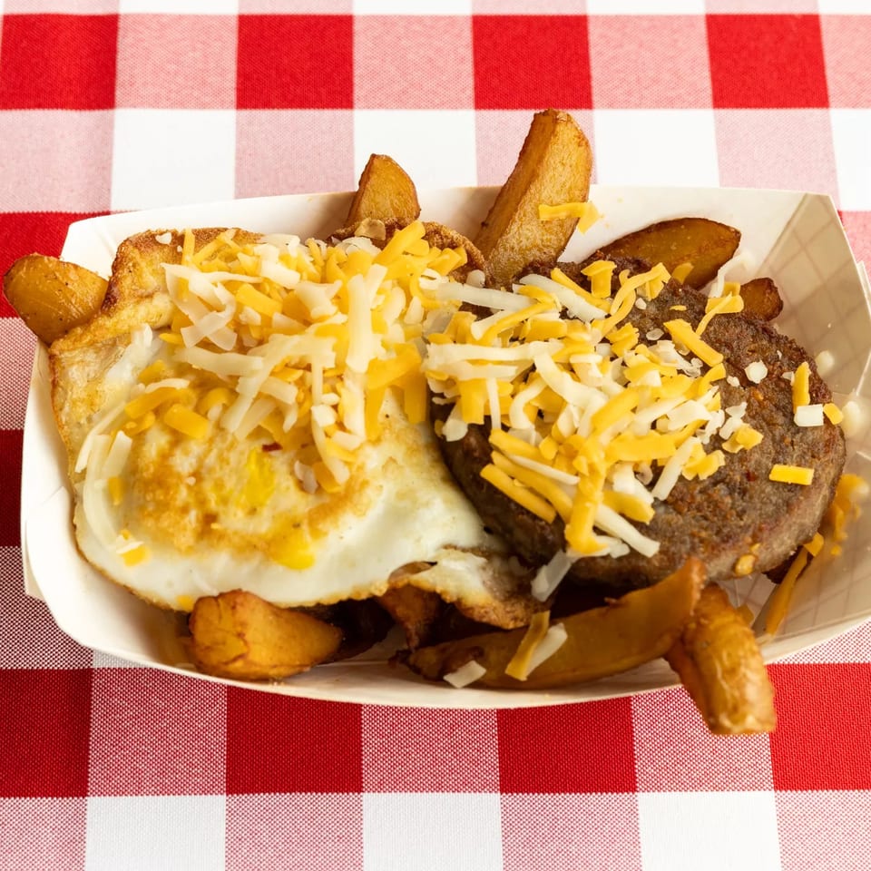 Breakfast platter with fried eggs, sausage patty, and potato wedges, all topped with shredded cheese.