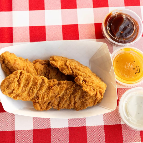 Crispy chicken tenders in a white tray with three dipping sauces on a red and white checkered tablecloth.