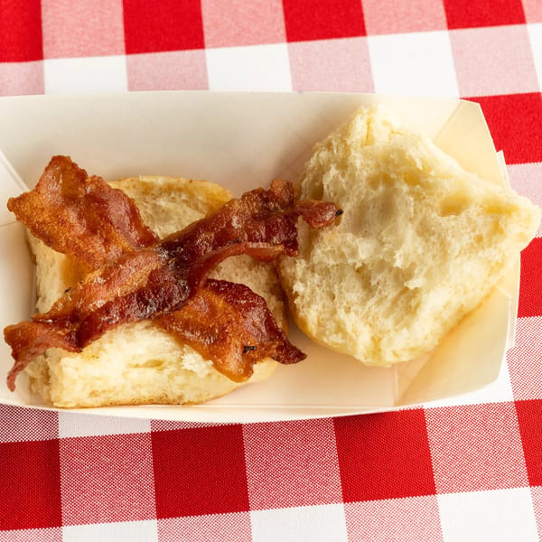 Two fluffy biscuits with crispy bacon strips in a white paper tray on a red and white checkered tablecloth.