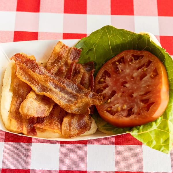 Crispy bacon strips, a large tomato slice, and lettuce on a white plate over a red and white checkered tablecloth.