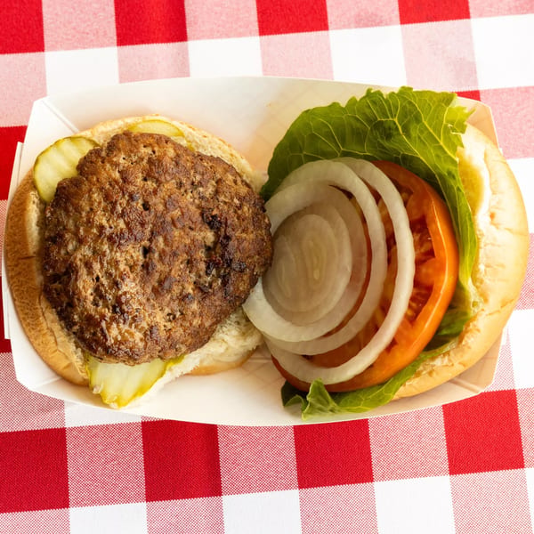 An open burger with a patty, pickles, lettuce, tomato, and onion on a red and white checkered tablecloth.