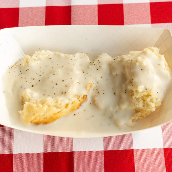 Biscuits and gravy in a white paper tray on a red and white checkered tablecloth.