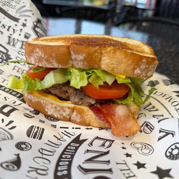A breakfast sandwich with bacon, lettuce, tomato, and cheese on toasted bread, served on branded paper.