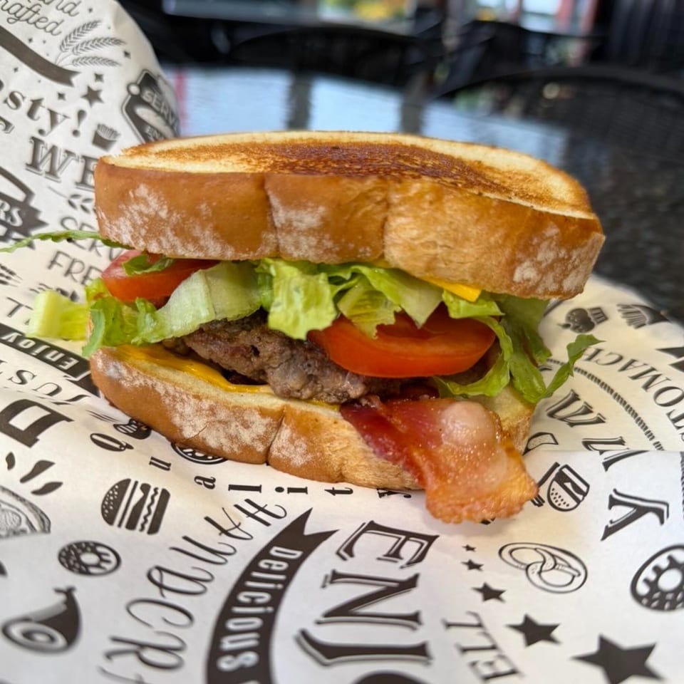 A breakfast sandwich with bacon, lettuce, tomato, and cheese on toasted bread, served on branded paper.