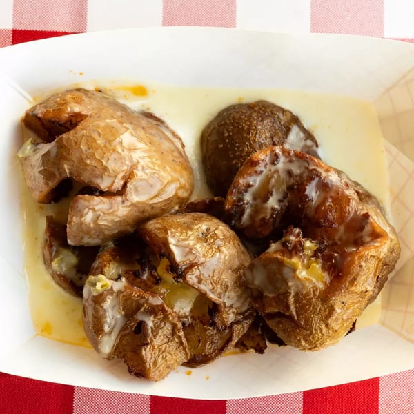 Crispy smashed potatoes drizzled with a creamy sauce, served in a white paper tray on a red and white checkered tablecloth.